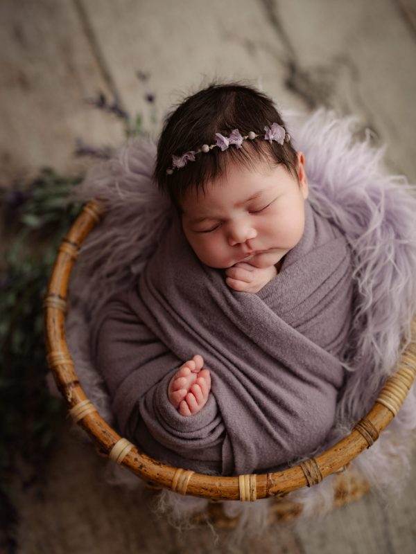 Sleeping newborn wrapped in a mauve blanket, wearing a flower headband, in a wicker basket.