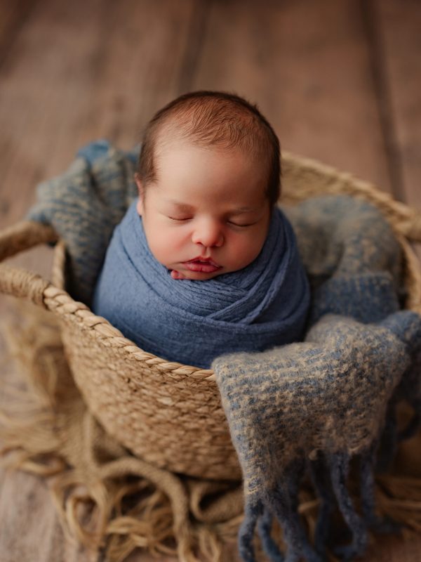 Newborn baby wrapped in a blue blanket, sleeping in a woven basket.