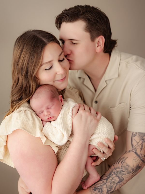 A mother holding a newborn baby while father gently kisses her cheek, all in cream clothing against a neutral background.