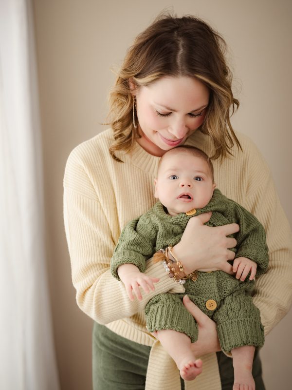 Mother gently holds a baby in a green knit outfit indoors, both looking down.